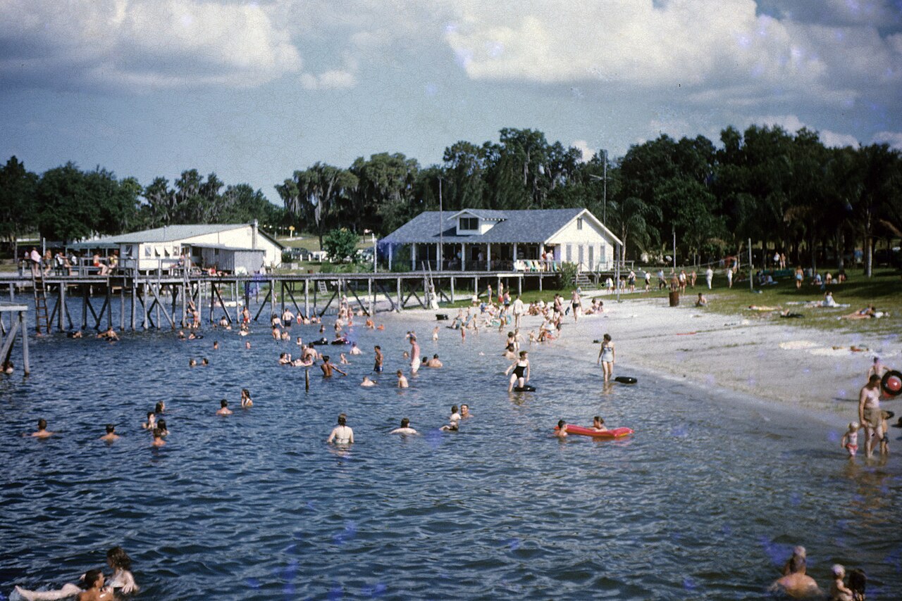 Swimmers in the shallows of a lake beach, with a pier and white pavilions behind, Ward’s Lake Iola, 1958