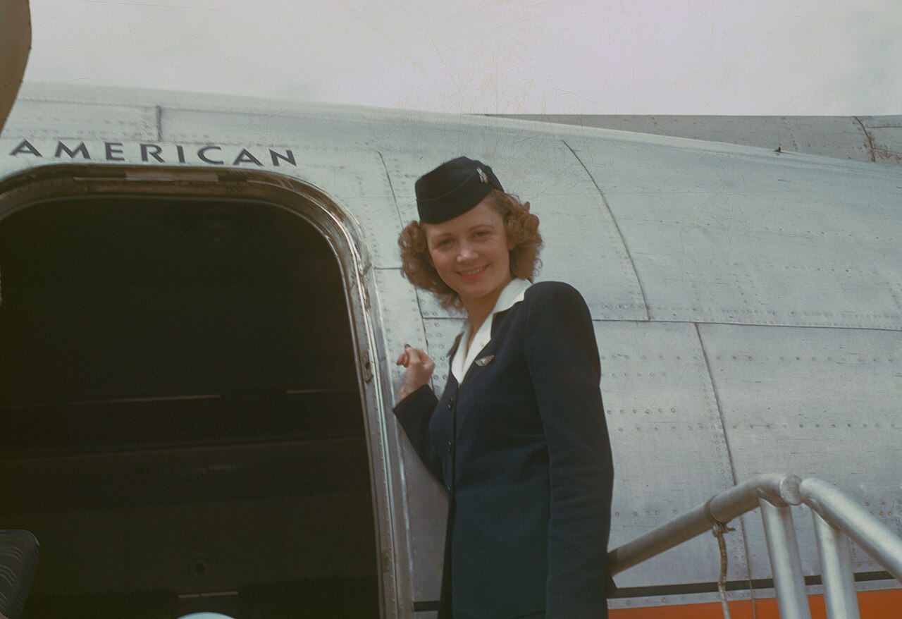 A stewardess in a dark uniform and cap smiles at the door of an American Overseas airliner, about 1949