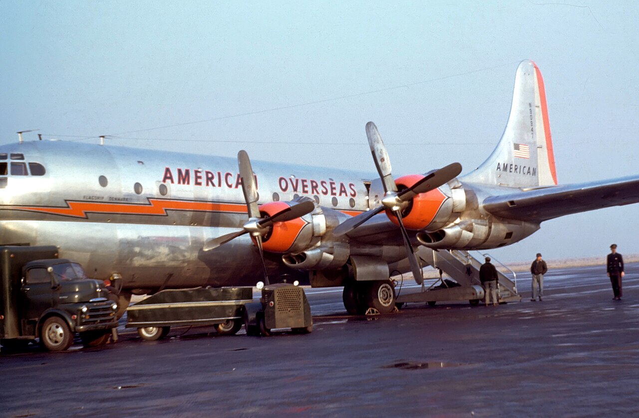 A silver American Overseas Boeing Stratocruiser with orange engine cowlings, parked on the apron, 1949–50